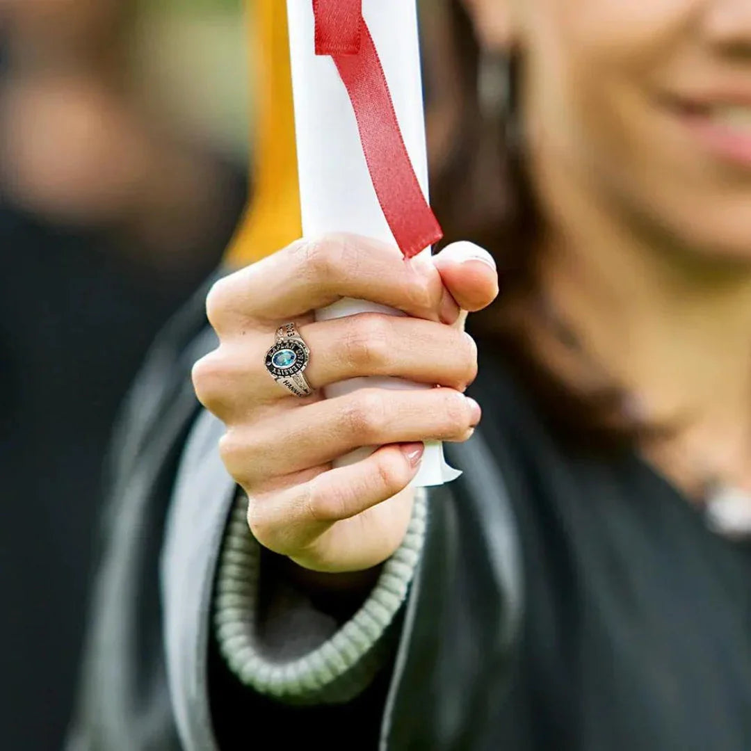 Graduate wearing personalized class ring with blue stone at graduation ceremony holding diploma - affordable and personalized class rings from Bella Olivia Gifts